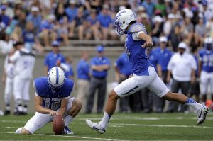 A football kicker in a blue and white uniform kicks the ball while another player holds it steady on the field. The kicker is mid-stride, wearing a helmet with a white stripe. Spectators and teammates in blue and white are visible in the background.