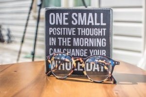 A pair of tortoiseshell glasses is resting on a wooden table in front of a small sign. The sign has a motivational quote written in white capital letters: "One small positive thought in the morning can change your whole day." The background, slightly blurred, adds an extra kicker to the scene.