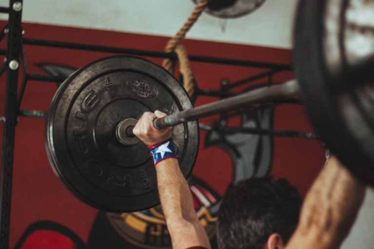 Close-up of a person lifting a heavy barbell in a gym, showcasing their resilience. The barbell has large weight plates, and the person's wrist, with a wristband featuring the Puerto Rican flag, is gripping the bar. A mural and various gym equipment are visible in the background.