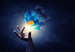 A hand reaches towards a glowing, floating 3D cube structure composed of smaller blue cubes, with one orange cube emitting a brighter light. The dark background is adorned with network-like lines and nodes, creating a futuristic and technological atmosphere that suggests complex problem-solving.