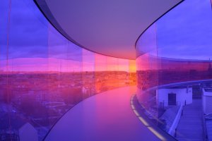 A curved corridor with transparent, tinted glass walls in shades of blue and pink offers a panoramic view of the cityscape at sunset. The colorful reflections and gradient effect create a surreal, vibrant atmosphere, evoking poetic words that dance through the scene.