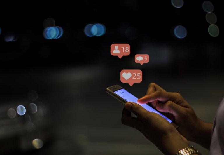 A person using a smartphone at night, with illuminated social media notification icons hovering above the screen. The notifications show 18 new followers, 5 direct messages, and 25 likes or reactions. The background is blurred with bokeh lights.