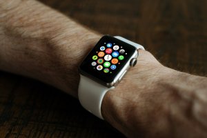 A close-up of a person's wrist wearing a smartwatch with a white band. The smartwatch screen displays colorful app icons, including social media apps, on a dark background. The person has a light skin tone and a hairy arm. The background is a dark wooden surface.