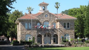 A historic stone building with a red-tiled roof, arched windows, a clock above the entrance, and multiple international flags displayed out front, surrounded by trees and a manicured lawn.