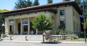 A historic yellow building with a red-tiled roof houses the Sonoma County Museum. The entrance features four large columns supporting a portico, and a banner is displayed above the steps. Lush greenery surrounds the building, and a small tree is prominent in the foreground.