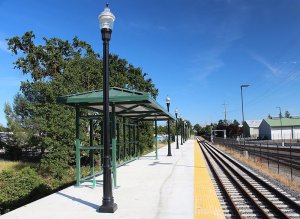 An empty train station platform with black lamp posts and green benches, sheltered by green roofs. The platform is bordered by a yellow safety line near the tracks. Trees and buildings are visible in the background under a clear blue sky.