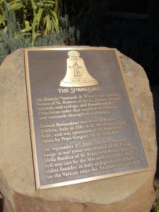 A plaque mounted on a rock commemorates the St. Francis Bell. Inscribed with information about St. Francis of Assisi, it notes his birth in 1181 and death in 1226. Images show a bell and the Vatican coat of arms. The plaque also details the bell's blessing in 2000.
