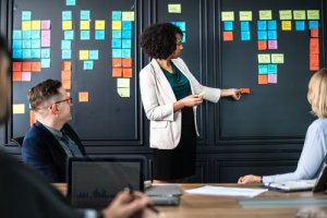 A woman with curly hair in a white blazer points at a wall filled with colorful sticky notes, leading a presentation on problem-solving. Two colleagues, one wearing glasses and a dark blazer and another with blonde hair, are seated at a table, listening attentively.