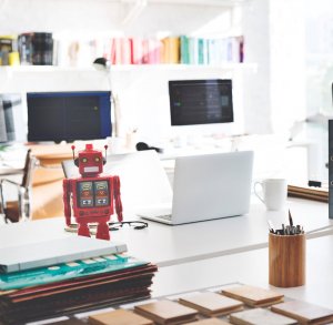 A brightly lit office space featuring a red toy robot on a desk, alongside a laptop, a cup, and stationery items. In the background, two computer monitors display artificial intelligence data, amid a stack of papers and shelves filled with colorful books.