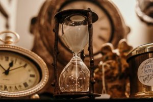 A close-up of an hourglass filled with white sand, surrounded by an assortment of antique clocks and decorative items. The hourglass is the central focus, with blurred background elements suggesting an antique or retro theme.