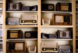 A wooden shelf filled with vintage radios of various shapes, sizes, and styles is adorned with both wooden and metal finishes. This collection of antiques showcases different models and designs, emphasizing a blend of retro aesthetics and historical technology.