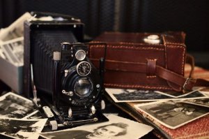 A vintage black camera is displayed on a surface surrounded by scattered black-and-white photographs. A brown leather case sits nearby, adding to the antique charm of the scene, evoking a nostalgic feel reminiscent of old memories and bygone eras of photography.