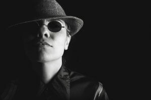 Black and white close-up of a person wearing round sunglasses and a fedora hat, casting a shadow over one side of their face. The person exudes a mysterious or noir vibe, with a serious facial expression, catching people’s attention in their dark outfit.