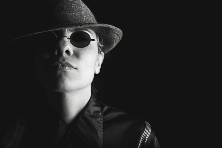 Black and white close-up of a person wearing round sunglasses and a fedora hat, casting a shadow over one side of their face. The person exudes a mysterious or noir vibe, with a serious facial expression, catching people’s attention in their dark outfit.