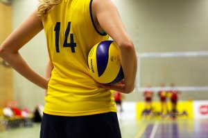 A volleyball player wearing a yellow jersey with the number 14 on the back is holding a volleyball. The player stands on a volleyball court, ready to catch people's attention with their skill. The net and other players are in the distance, while the slightly out-of-focus background shows an indoor gym setting.