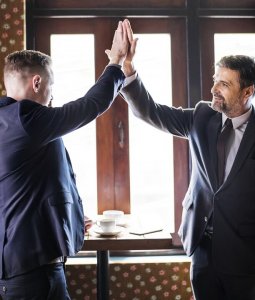 Two men in suits high-fiving in front of a window with natural light pouring in. They stand near a small table with coffee cups, looking pleased, as if they just managed to catch people’s attention during a successful business meeting.