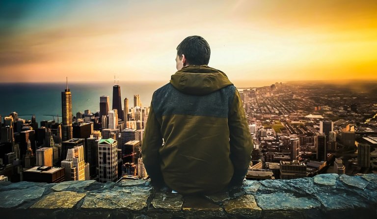 A person wearing a brown jacket sits on a stone wall, facing away and overlooking a cityscape at sunset. Skyscrapers and high-rise buildings stretch towards the glowing horizon, with a body of water visible to the left. The sky displays hues of orange and yellow, capturing the serenity akin to strong employee engagement.
