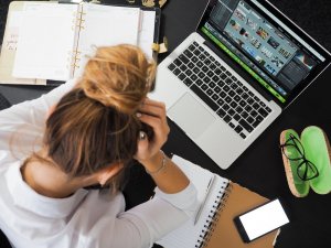 A person with long hair tied in a bun is sitting at a desk cluttered with a notebook, calendar, various papers, a smartphone, and a pen. They are holding their head in frustration while looking at a laptop displaying multiple images and graphics related to employee engagement.
