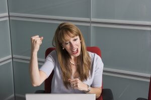 A woman with long hair and bangs sits in a red office chair, looking at a laptop screen. With a smile on her face and one arm raised with a clenched fist, she appears excited or victorious. In an office with frosted glass panels, her pose reflects high employee engagement.