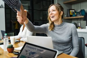 A woman with blonde hair, wearing a gray sweater, is seated at a wooden desk in an office. She is smiling and shaking hands with someone off-screen. In front of her is a laptop, and another laptop displaying a "Company's Growth" graph captures attention in the foreground.