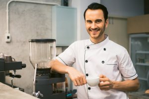 A smiling man in a white chef's jacket with black trim stands in a kitchen holding a coffee cup. He is leaning against a coffee grinder placed on the counter, drawing attention to the sleek appliances and well-organized shelves in the background.