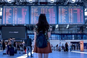 A woman with long hair and a backpack stands in front of a large digital departure board at an airport, contemplating a potential lateral transfer. She faces the board, which displays numerous flight schedules. Other travelers are seen in the background, some walking and others checking in.