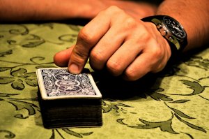 A person's hand, guided by consciousness, is seen pointing at or about to draw a card from a deck of playing cards resting on an ornate, green tablecloth. The person is wearing a dark wristwatch on their left wrist.