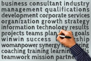 A person's hand holding a black marker writes on a light gray wall filled with various business-related words in bold font, including "business consultant," "strategy," "LinkedIn," "success," "teamwork," "goals," "growth," and "leadership.