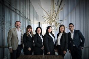 A group of six professionals, four women and two men, dressed in formal business attire, stand together indoors. They are posing for a LinkedIn-worthy photograph with a modern, artistic chandelier and floor-to-ceiling windows in the background. They all appear to be smiling.