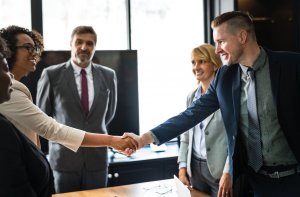 A group of four professionals are in a meeting room. One woman, wearing a beige jacket and eyeglasses, is shaking hands with a man in a suit and tie. Meanwhile, another man in a gray suit and a woman in a blue blouse and gray blazer are smiling in the background, possibly discussing resume length.