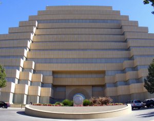 A large, tiered, beige building with a stepped, symmetrical design and horizontal lines. In front, there is a circular plaza with a seal and landscaped flower beds. Several cars are parked on either side.
