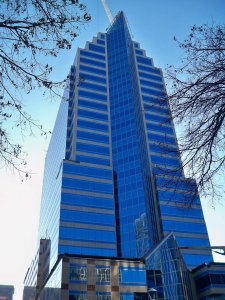 A tall, modern skyscraper with a blue glass facade and stepped design rises against a clear sky, framed by bare tree branches.