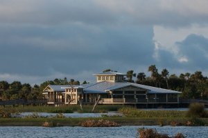 A modern building with a metal roof sits near a body of water, surrounded by palm trees and marsh vegetation under a cloudy sky.