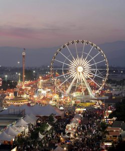 A lively Los Angeles County fairground at dusk features a large illuminated Ferris wheel, colorful rides, game booths, white tents, and crowds of people enjoying the event under a purple and pink sky.