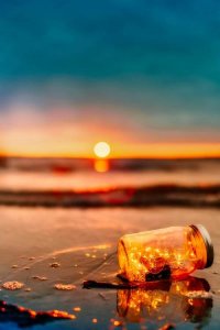 A glass jar filled with glowing fairy lights lies on wet sand near the ocean in Los Angeles County, with a vibrant orange sunset and calm water in the background.