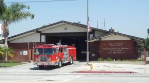 A red fire truck is parked outside a Los Angeles County brick fire station with a sign reading "City of Carson, Fire Station 127." An American flag is displayed in front.