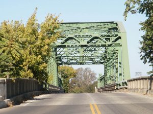 A green metal truss bridge with lattice framework crosses over a two-lane road, surrounded by trees on both sides under a clear sky.