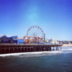 A large Ferris wheel and roller coaster sit atop a pier extending over the ocean, with buildings and palm trees visible in the background under a clear blue sky.