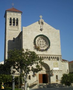 A beige stone church with a large circular window, a cross on top, a tall bell tower, arched wooden doors, and a wreath above the entrance, set against a clear blue sky with trees and steps in front.