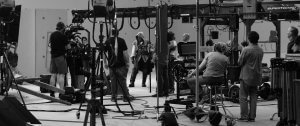 Black and white image showing a busy film set with various crew members operating cameras and sound equipment. Some are sitting, while others are standing at the front. Multiple pieces of filming equipment are visible, and actors in costume are preparing for a scene.