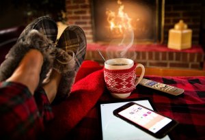 A cozy scene features someone's feet in slippers resting on a red blanket. A cup of steaming hot beverage in a red sweater mug sits next to a smartphone and tablet. In the background, a brick fireplace with a roaring fire adds warmth, accompanied by a TV remote ready for use.
