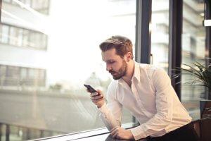 A man with a beard, wearing a white dress shirt, stands by a large window and looks intently at his smartphone. The cityscape outside is slightly blurred in the background, and there is a potted plant beside him. A cozy fireplace adds warmth to the sophisticated setting.