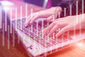 Hands typing on a laptop keyboard with overlaid line and bar graphs indicating data analysis or financial metrics, possibly in an Office 365 application. The background is slightly blurred and has a purple-pink hue.
