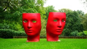 Two large, red, face-shaped sculptures with eye cut-outs are placed front and center on a grassy area outdoors. The faces have expressionless features and are surrounded by green trees and bushes, creating a striking contrast with the vibrant red color.