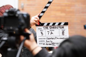 A film slate being held up front before a scene is shot. The slate shows details such as "Prod.: The Director," "Roll: 3," "Scene: 7A," "Take: 8," "Director: Henley Christopher," "Camera: Emily," and the date "05-26-2018." A person and a camera are visible in the foreground.