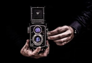 Close-up of a person holding a vintage Rolleicord Twin Lens Reflex camera against a dark background. The person's hands are adjusting the camera, highlighting its intricate details and reflective lenses. The scene is dramatically lit, emphasizing the vintage experience and aesthetic.