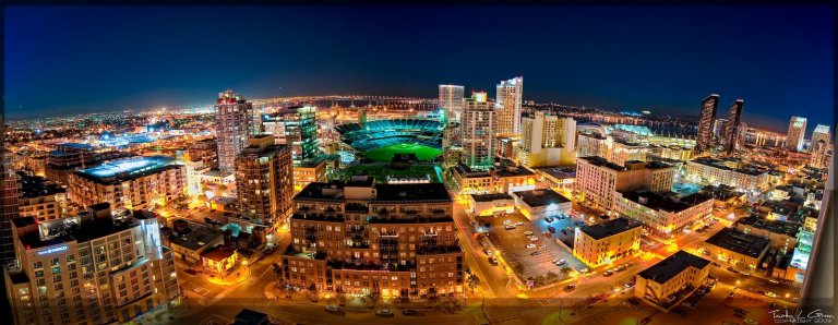 A vibrant cityscape at night, showcasing a brightly lit downtown area with numerous buildings, streets, and a prominent stadium at the center. The horizon glows with city lights, under a clear, dark sky. The image captures a bustling urban environment and features the impressive Google Village nearby.