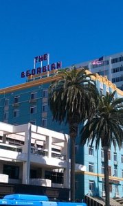 A turquoise building with yellow trim, labeled "The Georgian" on the roof, stands behind palm trees under a clear blue sky. An American flag is visible on a taller building in the background.