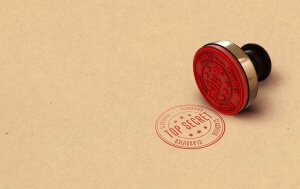 A red rubber stamp labeled "TOP SECRET" lies next to a stamped imprint of the same text on a brown, textured paper background. The secure imprint includes additional text around the border reading "CLASSIFIED" and "CONFIDENTIAL" along with small stars.