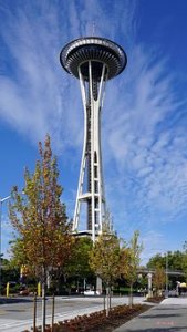 The Seattle Space Needle stands tall against a bright blue sky with scattered clouds, surrounded by green trees and a pedestrian walkway in the foreground, capturing the vibrant spirit of Seattle.